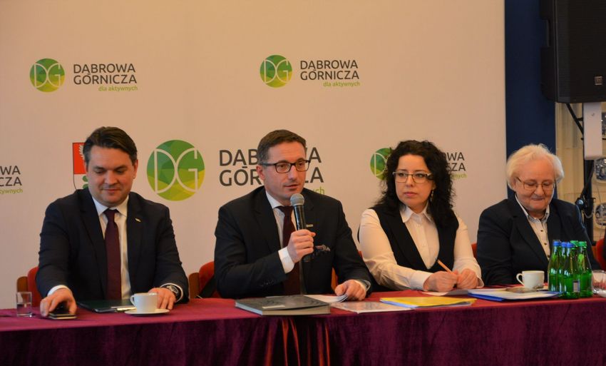 Cztery osoby uczestniczące w spotkaniu. Siedzą za stołem. Jedna osoba z mikrofonem.