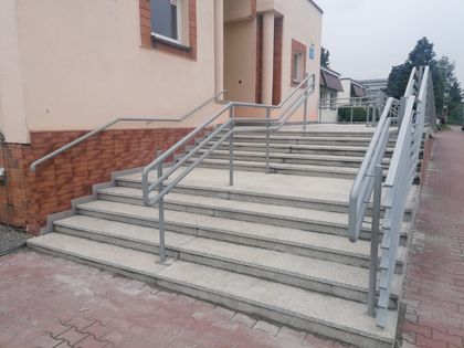 OsrodekJPII remont 15 Wyremontowane schody z podłożem lastrikowym i niklowanymi poręczami prowadzące do niskiego jasnego budynku. Obok chodnik z kostki betonowej.