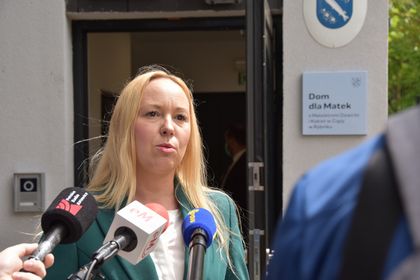 Kobieta w zielonej marynarce i białej bluzce na tle budynku. Przed nią mikrofony. 