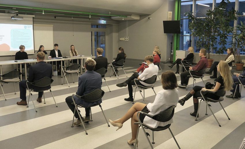 Młodzież siedząca na pojedynczych krzesłach, rozmieszczonych w odstępach na sali 
