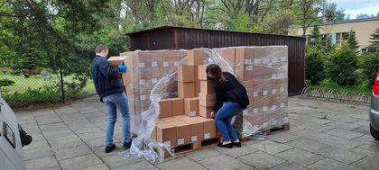 Maseczki fot 4 Dwie osoby, kobieta i mężczyzna, rozładowują paczki na podjeździe. W tle zieleń.
