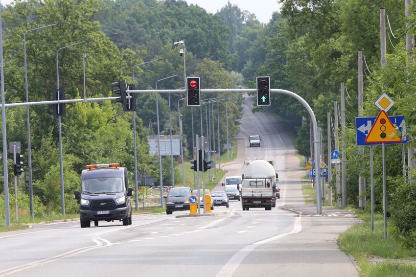 Droga dwukierunkowa otoczona lasem, jadące nią samochody. Nad drogą sygnalizacja świetlna