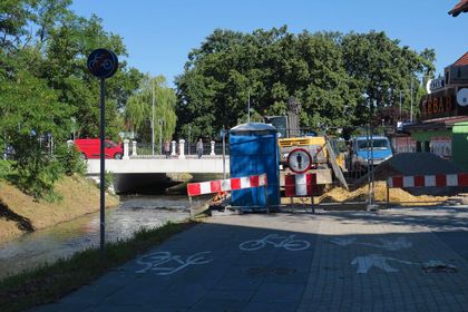 Roboty budowlane na drodze wzdłuż rzeki. W tle most, wokół drzewa i zabudowa miejska. 