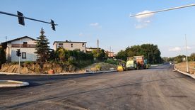 Droga w trakcie remontu, świeżo położony asfalt. Wzdłuż drogi domy, zieleń.