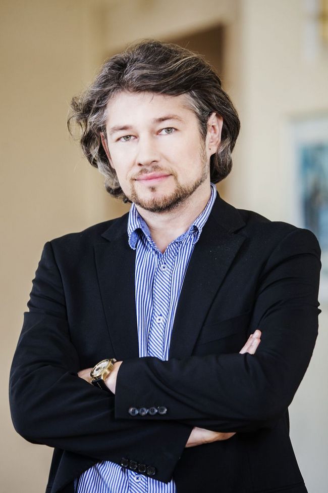 Mężczyzna stojący z założonymi rękami, w ciemnym garniturze.