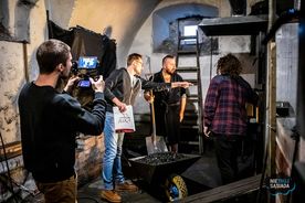 Cztery osoby stoją w kotłowni obok taczki z węglem. Mężczyzna na pierwszym planie trzyma w reku kamerę. 