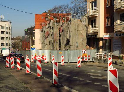 Remont kamienicy. Fasada budynku zasłonięta płachtami. Przed budynkiem blaszany płot. Na pierwszym planie ulica i pachołki wyznaczające nową organizację ruchu. 