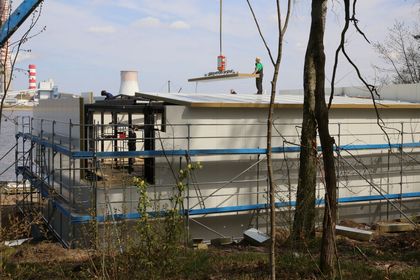Budynek kontenerowy otoczony rusztowaniem w trakcie rozbudowy. W tle niebo, zalew i kominy elektrowni.