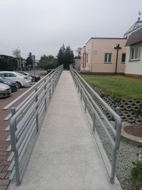 OsrodekJPII remont 19 Podjazd dla osób niepełnosprawnych. W tle zabudowania i szare niebo, po lewej stronie zaparkowane samochody, po prawej trawnik.