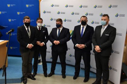 Pięciu mężczyzn w ciemnych garniturach i maseczkach na twarzy w trakcie konferencji prasowej. W tle banery reklamowe miasta Rybnika i subregionu zachodniego. 