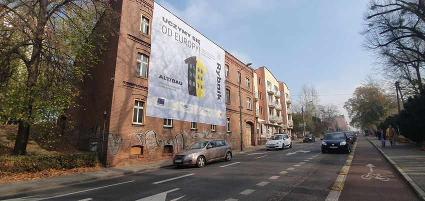 Ulica, po jej lewej stronie ceglana kamienica, częściowo zasłonięta materiałowym bannerem. 
