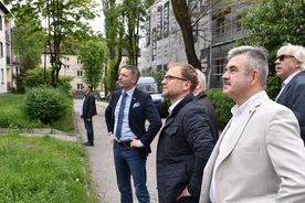 Grupa elegancko ubranych mężczyzn patrzy na coś przed siebie. W tle zieleń i zabudowania.