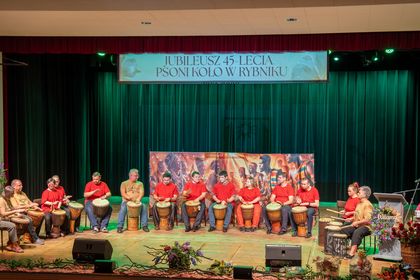 Grupa osób siedzi na scenie i uderza w bębny.