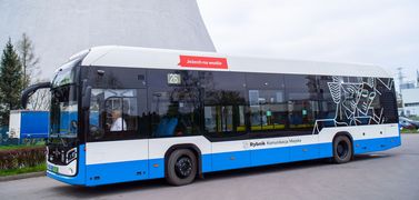 Na zdjęciu widać autobus w całości, bokiem. W tle komin elektrowni.