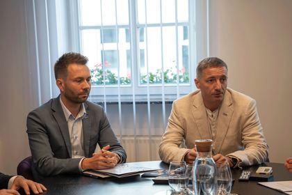 Zdjęcie przedstawia dwóch mężczyzn siedzących przy stole na oficjalnym spotkaniu.