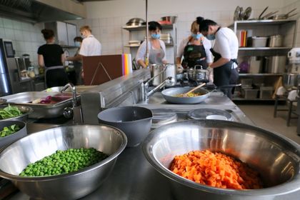 Różnokolorowe produkty spożywcze na kuchennym blacie, w tle osoby pracujące przy przygotowywaniu posiłków.