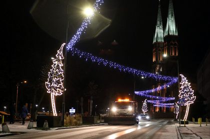 Pługopiaskarka odśnieżająca ulicę. W tle oświtlony kościół.