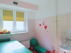 Pokój z materiałami do rehabilitacji. Czerwony materac, zielona piłka i wałek. Okno z żółtą roletą, zlew. Na jasnych ścianach przyklejanki - kosz z balonem i chmury. 