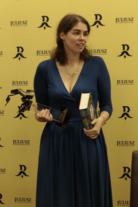 Kobieta w granatowej sukience ze statuetką i książką w ręku na tle żółtej ścianki promocyjnej nagrody Juliusz.