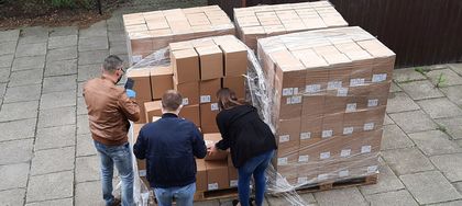 Maseczki fot 3 Trzy osoby, kobieta i 2 mężczyzn, rozładowują paczki na podjeździe. W tle zieleń za płotem i blaszany garaż.