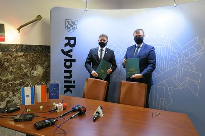 Dwóch mężczyzn stojących na tle ścianki z napisem Rybnik. Obaj panowie trzymają w rękach zielone, papierowe teczki. 