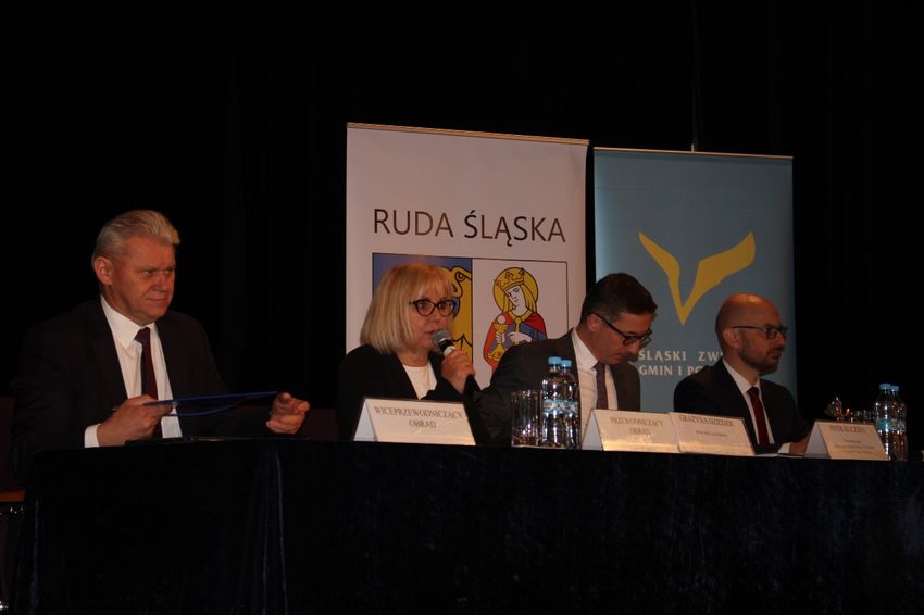 Cztery osoby, w tym jedna kobieta, siedzą za stołem konferencyjnym, w tle banery reklamowe. 