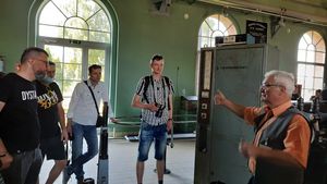 Kilku młodych mężczyn slucha wykładu starszego mężczyzny w sali z maszyną parową