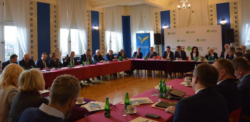 Grupa osób siedzących przy stołach wokół sali narad. 