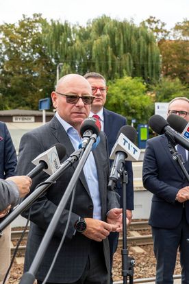 Zdjęcie przedstawia mężczyznę mówiącego do mikrofonu. W tle widać dwóch mężczyzn.