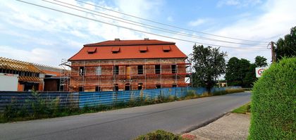 Piętrowy, ceglany budynek pocysterskiego dworu w trakcie remontu. Widoczne rusztowania. Obok inne zabudowania w trakcie budowy. Na pierwszym planie droga, w tle błękitne niebo z białymi chmurami. 