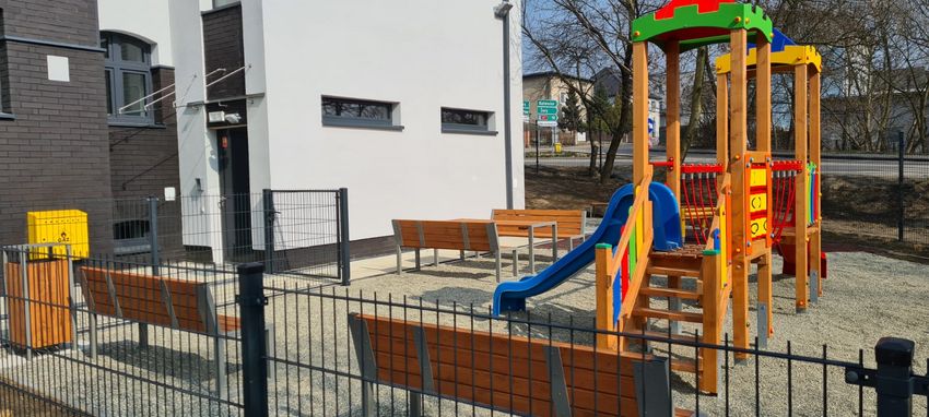 Biały, dwupiętorowy budynek otoczony płotem, za płotem plac zabaw