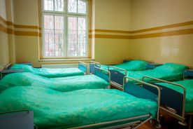 Jasna sala szpitalna z oknem, po obu stronach rząd zasłanych łóżek z zieloną pościelą 