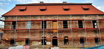Piętrowy, ceglany budynek pocysterskiego dworu w trakcie remontu. Widoczne rusztowania, w tle błękitne niebo z białymi chmurami. 