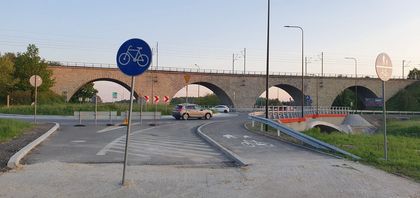 Fragment ulicy i drogi rowerowej. W tle wiadukt. Na pierwszym planie znak drogowy - rower w okręgu na niebieskim tle. 