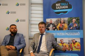 Dwaj mężczyźni siedzą na tle banerów reklamowych. 