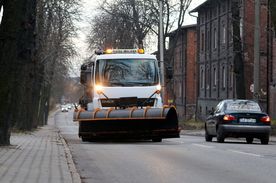Ulice miasta, na nich pługopiaskarka i pracownicy służb komunalnych z szuflami do odśnieżania.