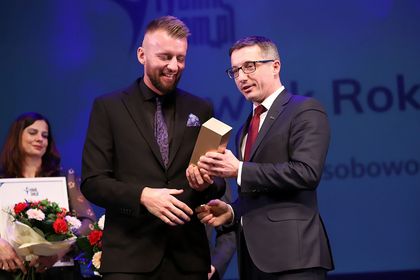 Mężczyzna przekazuje statuetkę drugiemu mężczyźnie. W tle kobieta z dyplomem. 