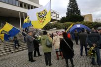 Kilka osób na otwartej przestrzeni, przy schodach prowadzących do teatru. Ubrani na zimowo ludzie trzymają w rękach flagi śląskich organizacji. 