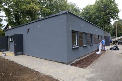 Niski szary budynek, po prawej stronie dwóch mężczyzn w trakcie prac budowlanych i porządkowych, w tle zieleń.