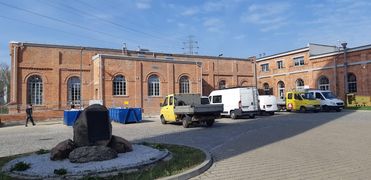 Zmodernizowane zabudowania byłej kopalni - budynek z cegły na tle błękitnego nieba. Na pierwszym planie zaparkowane samochody i obelisk. 