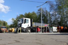 Samochód ciężarowy tankujący paliwo na stacji CNG. W tle inne samochody i zieleń. Na pierwszym planie betonowy plac.