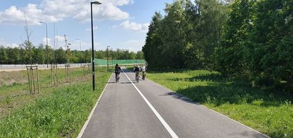 Trzy rowerzystki widziane z tyłu i z oddali na drodze rowerowej. Po obu stronach zieleń. W dali, po lewej stronie, ekrany akustyczne i barierki przy drodze szybkiego ruchu. 