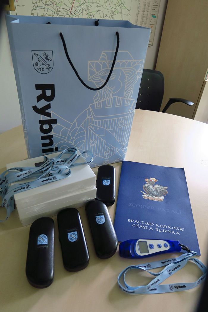 Przedmioty leżące na stole: termometr medyczny, cztery czarne etui z herbem miasta, cztery smycze z nadrukiem Rybnik, w tle torba papierowa Rybnik oraz teczka Bractwo Kurkowe.