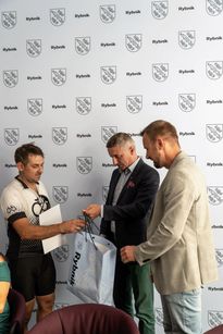Zdjęcie przedstawia trzech mężczyzn. Dwóch z nich wręcza prezent i dyplom trzeciemu.