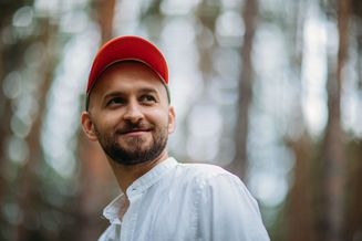 Mężczyzna z małym zarostem, w jasnej koszuli i czerwonej czapce z daszkiem, pokazany od pasa w górę na tle rozmytej zieleni - artysta Baranovski