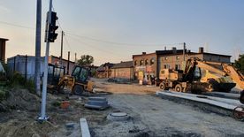 Droga w trakcie remontu, świeżo położony asfalt. Wzdłuż drogi domy, zieleń.