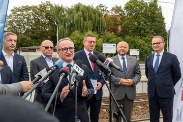 Zdjęcie przedstawia grupę mężczyzn stojących przy mikrofonach na peronie pkp. Jeden z mężczyzn mówi do mikrofonu.