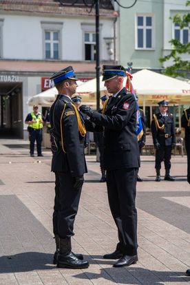 Zdjęcie przedstawia dwóch mężczyzn w mundurach strażackich. W tle ludzie.