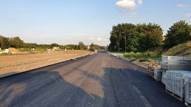 Droga w trakcie remontu, świeżo położony asfalt. Wzdłuż drogi domy, zieleń.