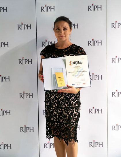 Kobieta w ciemnej sukience trzyma dyplom na tle ścianki promocyjnej Regionalnej Izby Przemysłowo-Handlowej